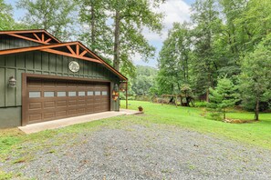A rustic cabin with a garage, surrounded by a well-maintained lawn and dense forest. - Deck & Game Room! Lakefront Blue Ridge Mtn Gem (Deep Gap)