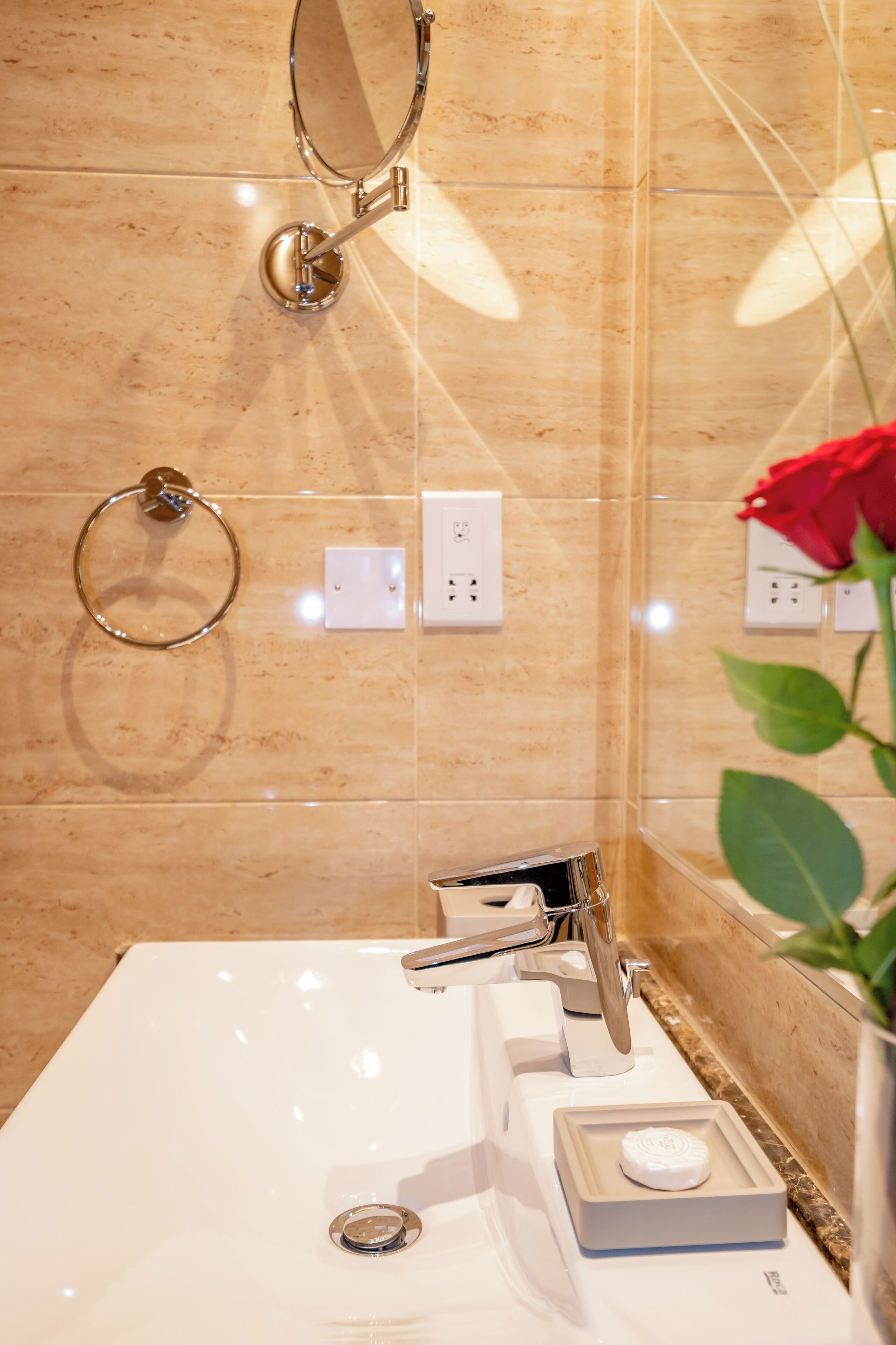 A bathroom with a white sink, a mirror, a towel ring, and a red rose in a vase.