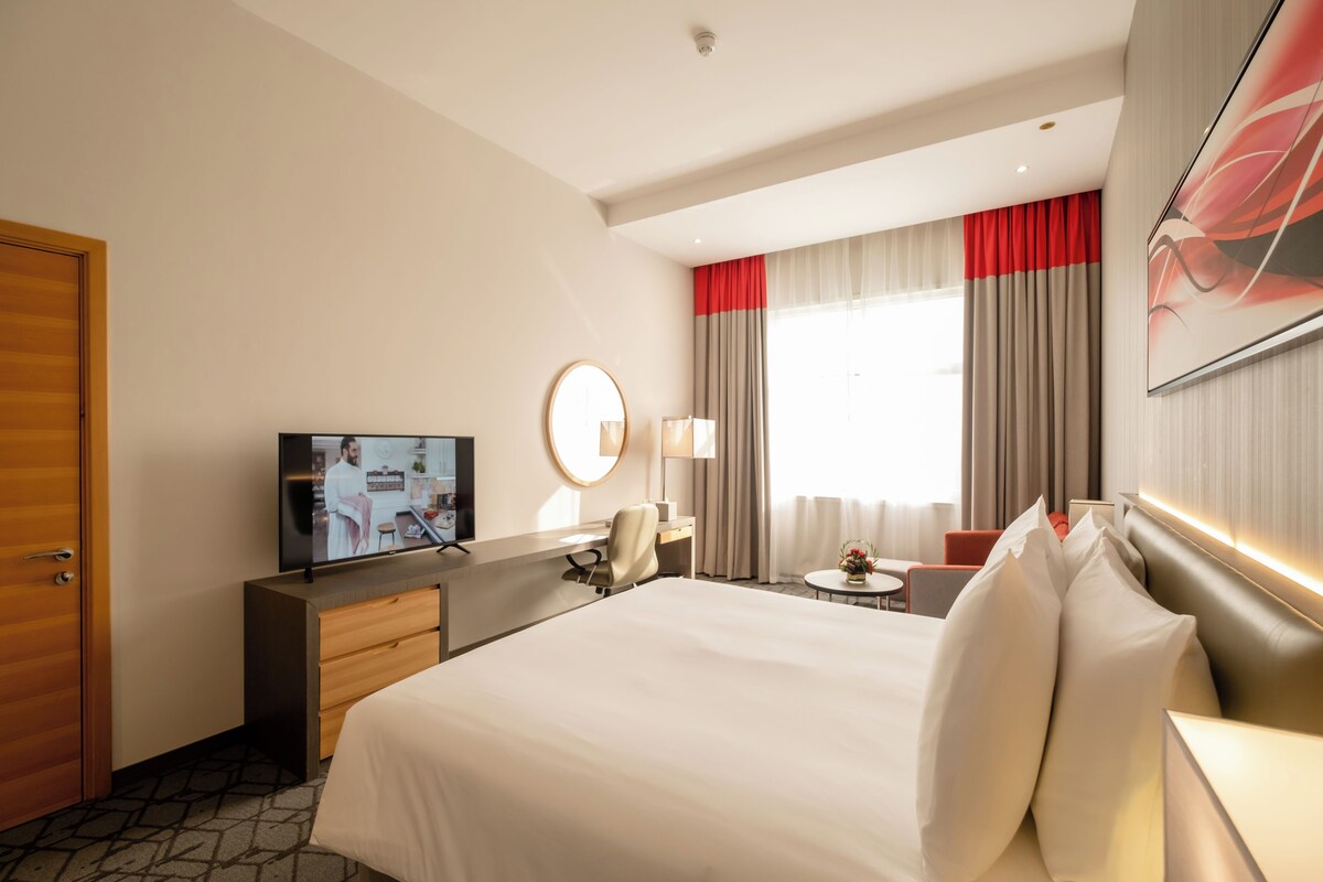 Quarto de hotel com uma cama grande, uma escrivaninha com cadeira, televisão, espelho redondo, mesinha de cabeceira com abajur e janela com cortinas.