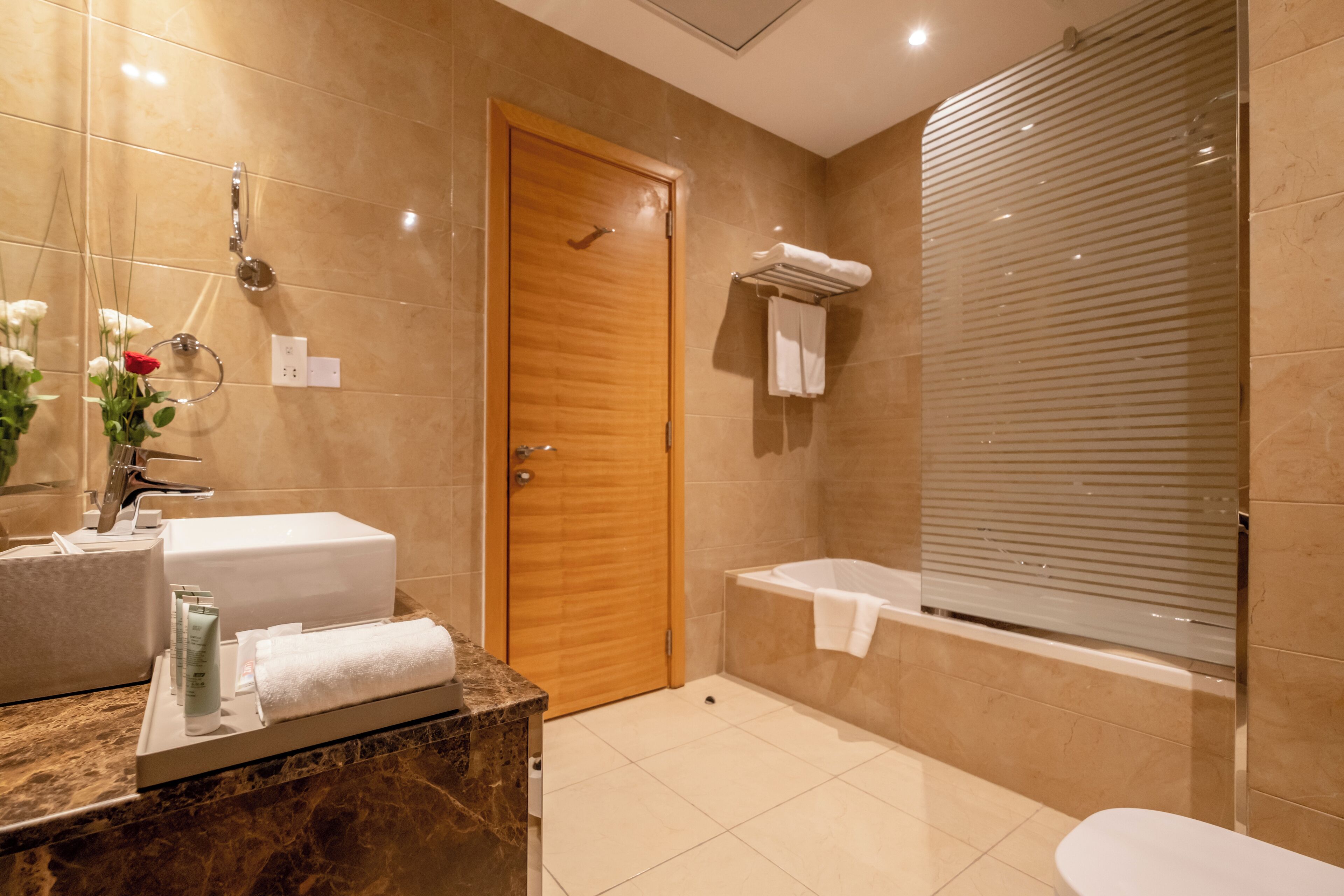 A modern bathroom with a wooden door, a bathtub, a shower, and a marble countertop with toiletries.
