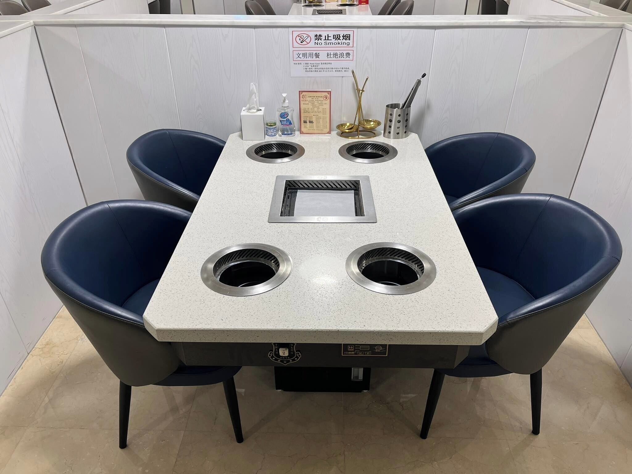 A table with a built-in grill, four blue chairs, a no-smoking sign, and a menu on the wall.