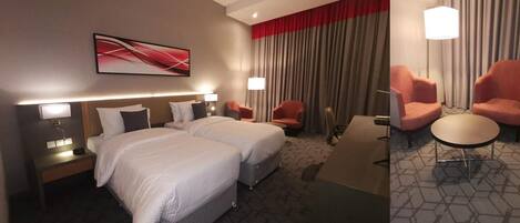 A hotel room with two beds, a desk, two armchairs, and a large window with curtains.