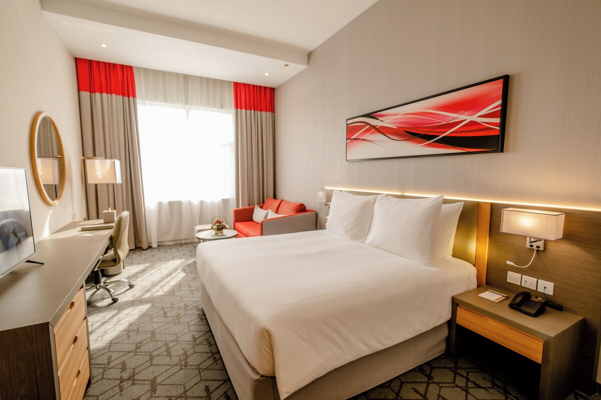 A hotel room with a large bed, a desk with a chair, a TV, and a desk lamp.