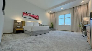 Habitación de hotel con cama, mesitas de noche, escritorio, silla, ventana grande y un cuadro en la pared.