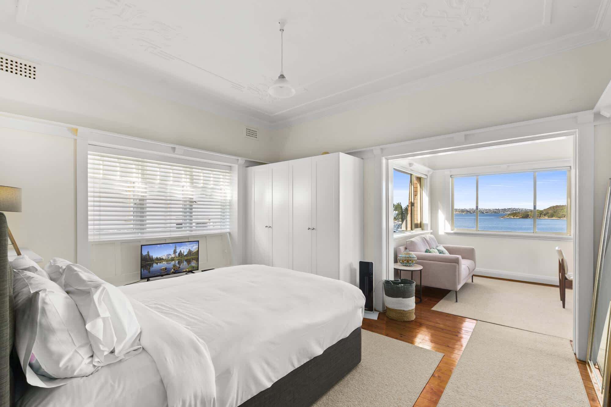 A bedroom with a bed, a TV, a wardrobe, a desk, a chair, and a view of the sea.