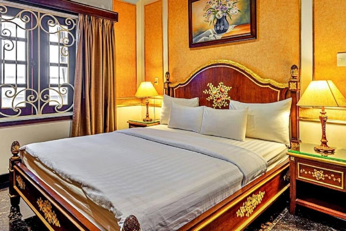 Ein Hotelzimmer mit einem großen Bett, einem aufwendig geschnitzten Holzkopfstück, zwei Nachttischen mit Lampen, einem Wandbild und einem Fenster mit Vorhängen.