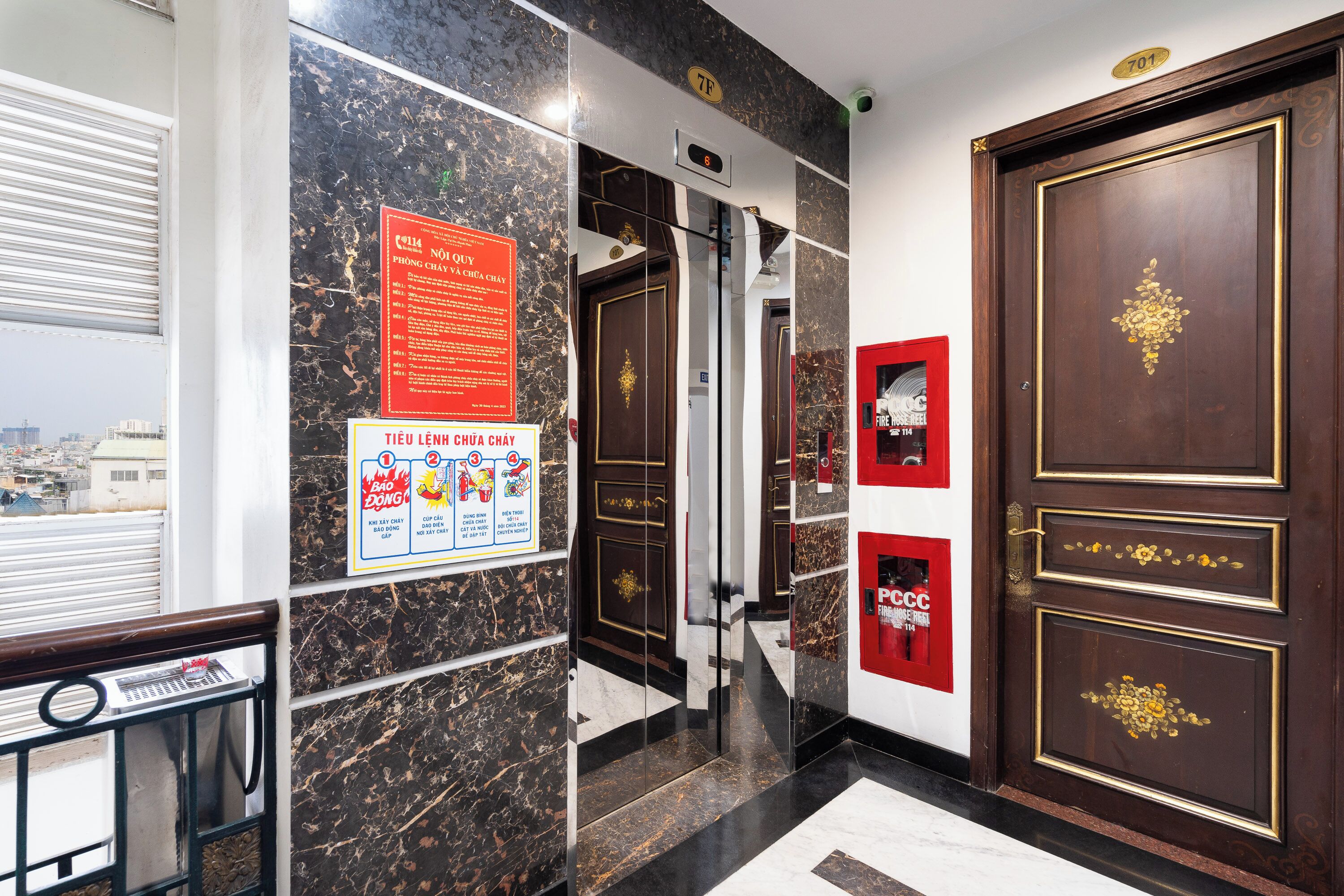 Un vestíbulo de hotel con piso de mármol, un cartel de seguridad contra incendios, un panel digital de ascensor y una puerta con manguera contra incendios.