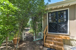 A wooden staircase leading to a dark-colored door with a window, surrounded by greenery and a white picket fence.