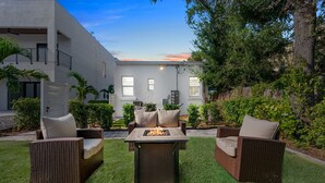 A patio with wicker furniture and a fire pit, surrounded by a well-maintained lawn and a modern building in the background. - Pineapple Paradise #1 • 13 min to Beach • Fire Pit (Sarasota)