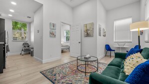 A modern living room with a green sofa, a coffee table, and a colorful rug. There is a dining area with a bench and a table, and a kitchen area with a refrigerator and a chair.