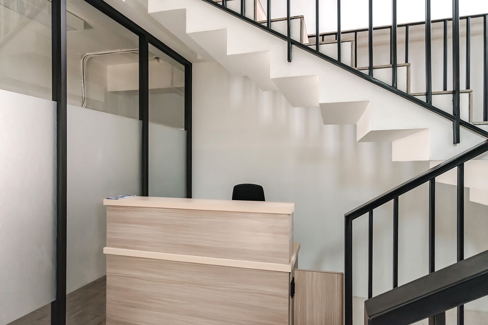 A modern reception desk with a light wood finish, a black chair, and a staircase with black railings.