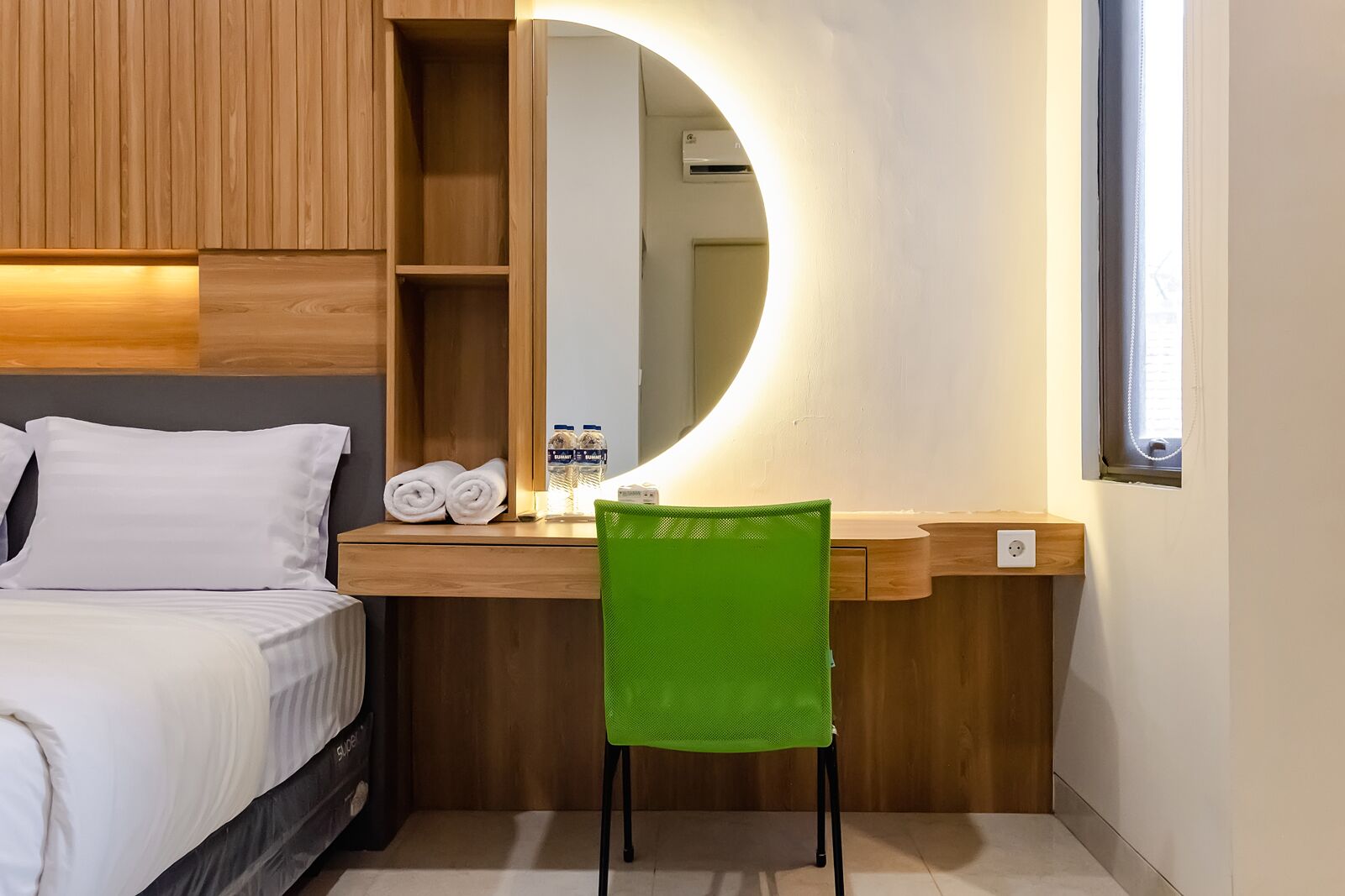 A hotel room with a bed, a desk with a mirror, a green chair, and a small window.