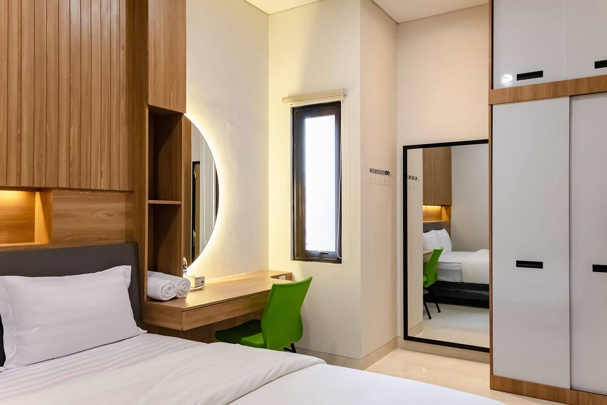 A hotel room with a bed, a desk with a mirror, a green chair, and a wardrobe.