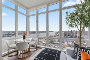 Una habitación amplia con ventanas grandes que ofrecen vista a la ciudad, una mesa redonda con sillas y un sofá con almohadones decorativos.
