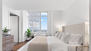 Une chambre avec un grand lit, une commode, une fenêtre donnant sur des immeubles et une plante en pot.