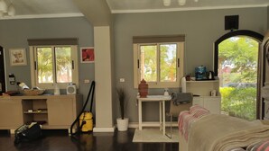 A living room with a sofa, a coffee table with a cup, a vacuum cleaner, a window with a view of trees, and a small table with a plant. - Villa 56 re (October Gardens)