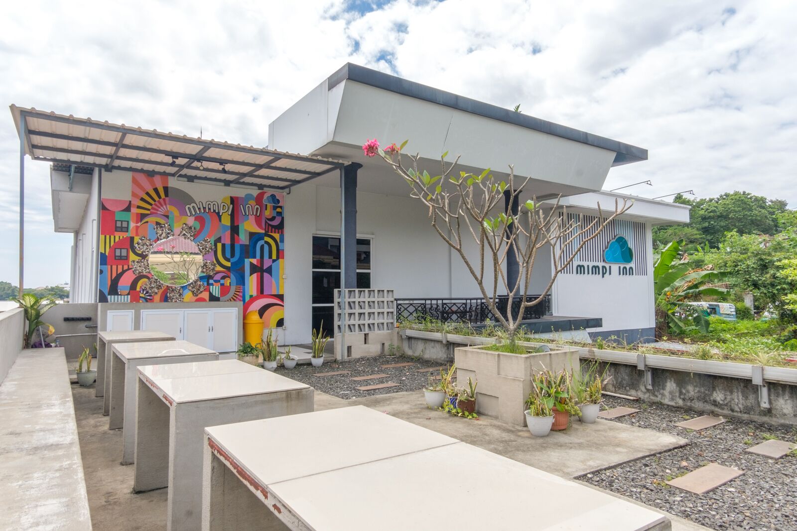 A modern building with a colorful mural on the side, outdoor seating, and a small tree in a planter.
