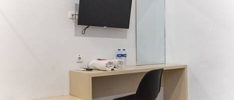 Quarto moderno com escrivaninha de madeira, cadeira preta, televisão de tela plana e um espelho grande. Há duas garrafas de água e uma toalha enrolada na escrivaninha.