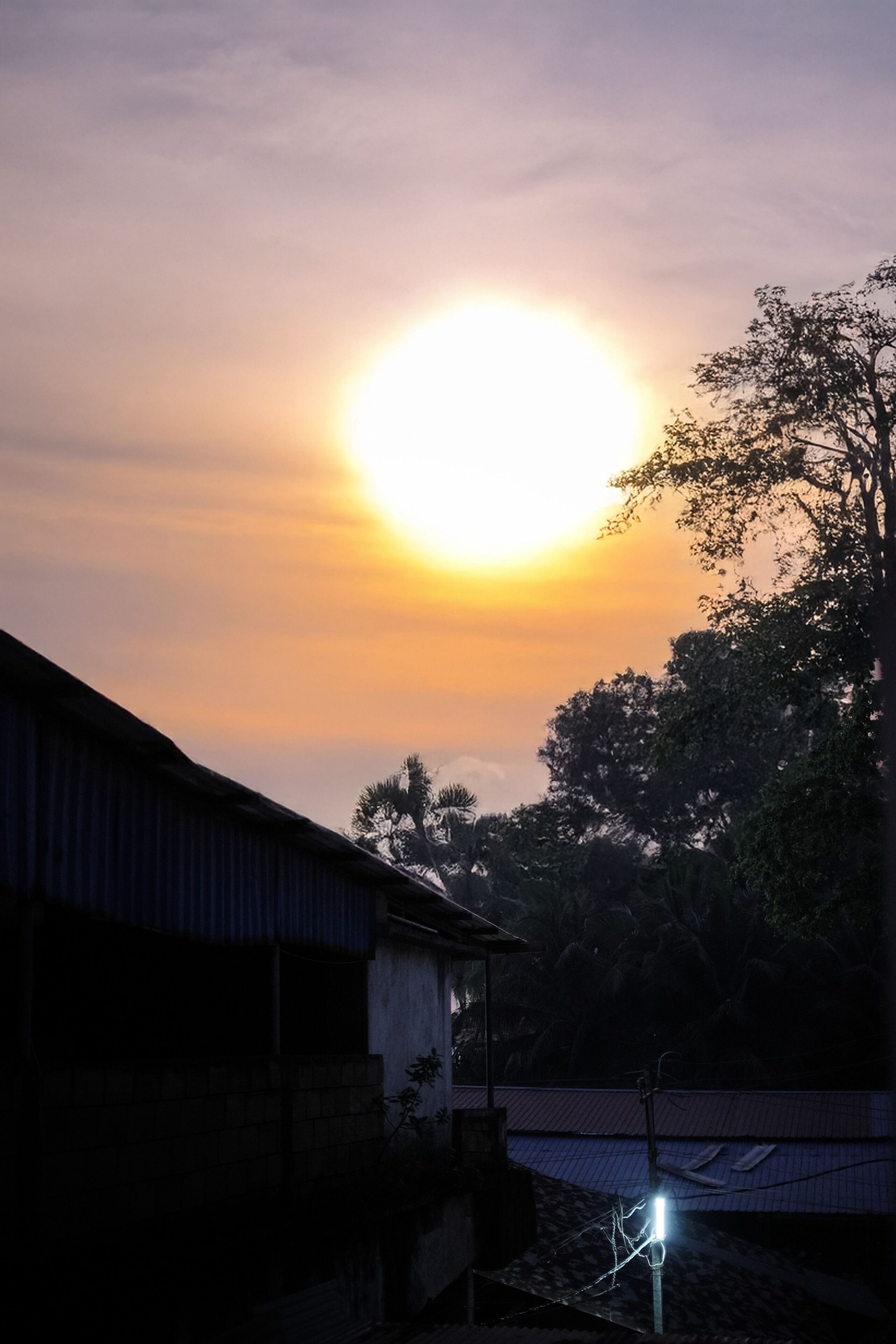 A sunset with a bright sun, a building, and trees in the background.