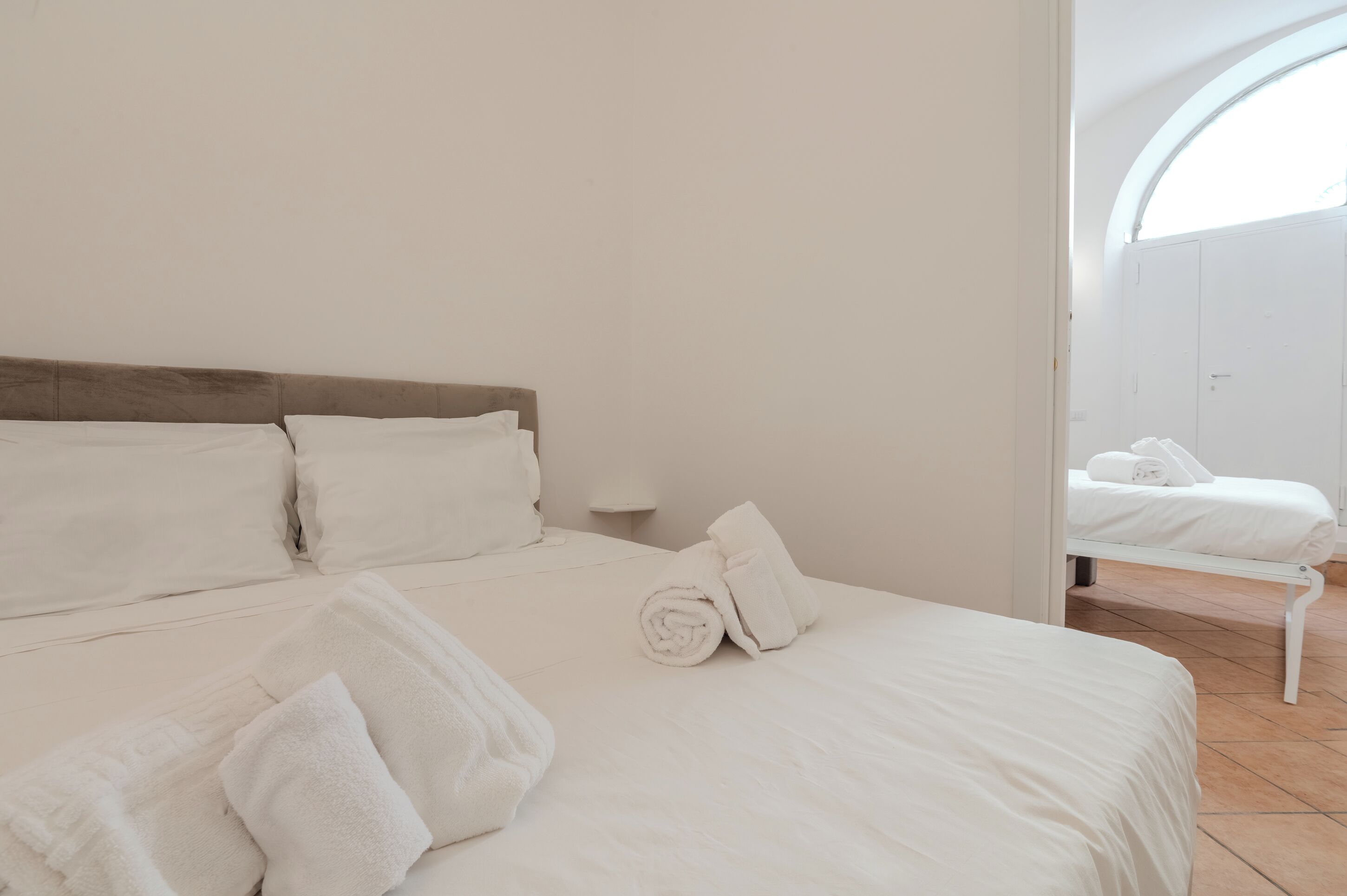 A clean, white-themed bedroom with a neatly made bed, a small bedside table, and a view through an arched window.