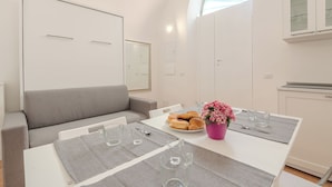 A modern kitchen with white cabinets, a dining table set with placemats, a plate of croissants, a pink flower vase, and glassware.
