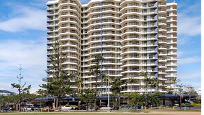 A multi-story hotel with balconies, a parking area, and palm trees. - 26Dec 2026 to 2Jan 2027 Beach House Accommodation - New Years (Coolangatta)