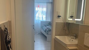 A modern bathroom with a white sink, mirror, and toilet. There is a towel rack with towels on the wall. The bathroom leads to a bedroom with a bed and a window with curtains.