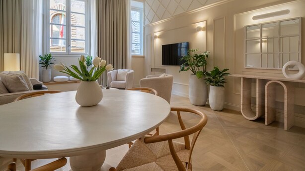 Una sala de estar moderna con una mesa redonda, asientos cómodos, un televisor de pantalla plana montado en la pared y ventanas grandes con cortinas.