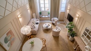 Una sala de estar moderna con un sofá, sillones, una mesa de centro y una televisión. Hay plantas en macetas y un cuadro en las paredes.