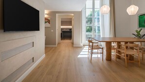 Ein modernes Wohnzimmer mit einem an der Wand befestigten Flachbildfernseher, einem Essbereich mit Holzmöbeln und einer durchgehenden Tür erreichbaren Küche.