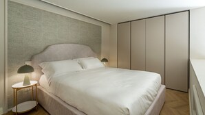 A modern bedroom with a large bed, a headboard with a textured pattern, a bedside table with a lamp, and a wardrobe with vertical doors.