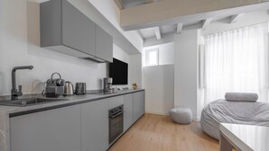 Une cuisine moderne équipée d’une cuisinière intégrée, d’un évier et d’une machine à café. Une grande fenêtre avec des rideaux légers et un lit recouvert d’une couverture grise se trouvent dans la pièce adjacente.