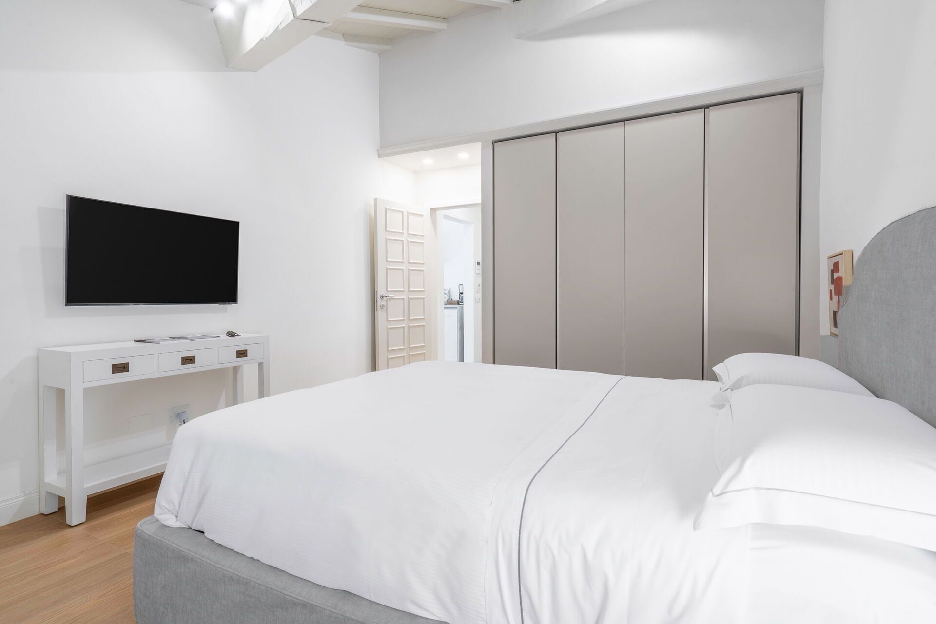 A modern bedroom with a large bed, a TV mounted on the wall, a white dresser, and a wardrobe.