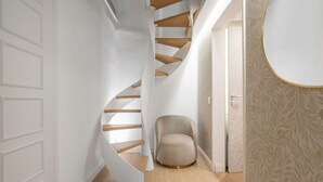 Een wenteltrap met houten treden en een beige fauteuil in een hal met behang met een patroon en een witte deur.