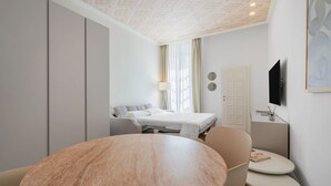 Une chambre d’hôtel moderne avec une table à manger ronde, un canapé, un lit avec une tête de lit blanche, une table de chevet avec une lampe, un téléviseur fixé au mur et une porte avec un miroir.