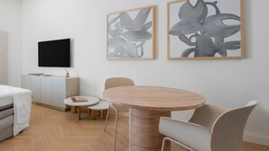 Un salon moderne avec une table ronde en bois, deux chaises, un téléviseur à écran plat fixé au mur et des tableaux abstraits représentant des motifs floraux.