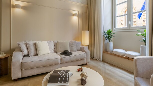 Un salon moderne avec un canapé beige, une table basse ronde et une lampe. On peut voir deux plantes en pot sur un rebord de fenêtre, et une vue sur un bâtiment avec un drapeau à l’extérieur.