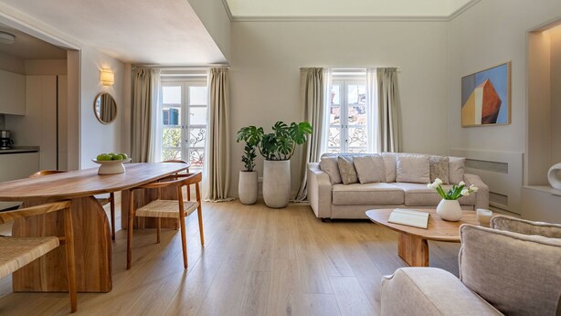 Een moderne woonkamer met een houten eettafel, een beige bank en een salontafel. Er staan potplanten en er hangt een schilderij aan de muur.