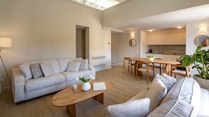 Een ruime woonkamer met een lichtkleurige bank, een houten salontafel en een eettafel met stoelen. In de achtergrond is een keuken met witte kastjes en een marmeren achterwand zichtbaar.