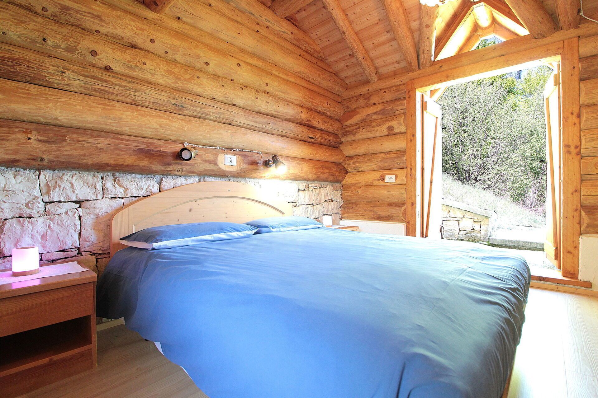 Camera da letto in una baita di legno con parete in pietra, un letto, un comodino e una finestra con vista su vegetazione.