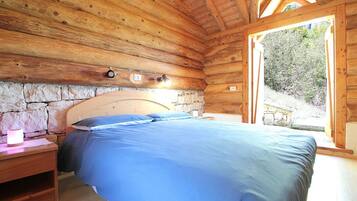 Camera da letto in una baita di legno con parete in pietra, un letto, un comodino e una finestra con vista su vegetazione.
