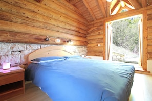 A wooden cabin bedroom with a stone wall, a bed, a nightstand, and a window with a view of greenery.