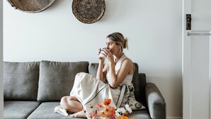 A person sitting on a grey sofa with a white cushion, holding a cup. There is a wooden tray with a book and a wallet on a side table, and a vase with flowers on the floor. - Benambra Boutique Accommodation (Queenscliff)