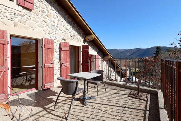 A stone building with red shutters, a balcony with a table and chairs, and a view of mountains.