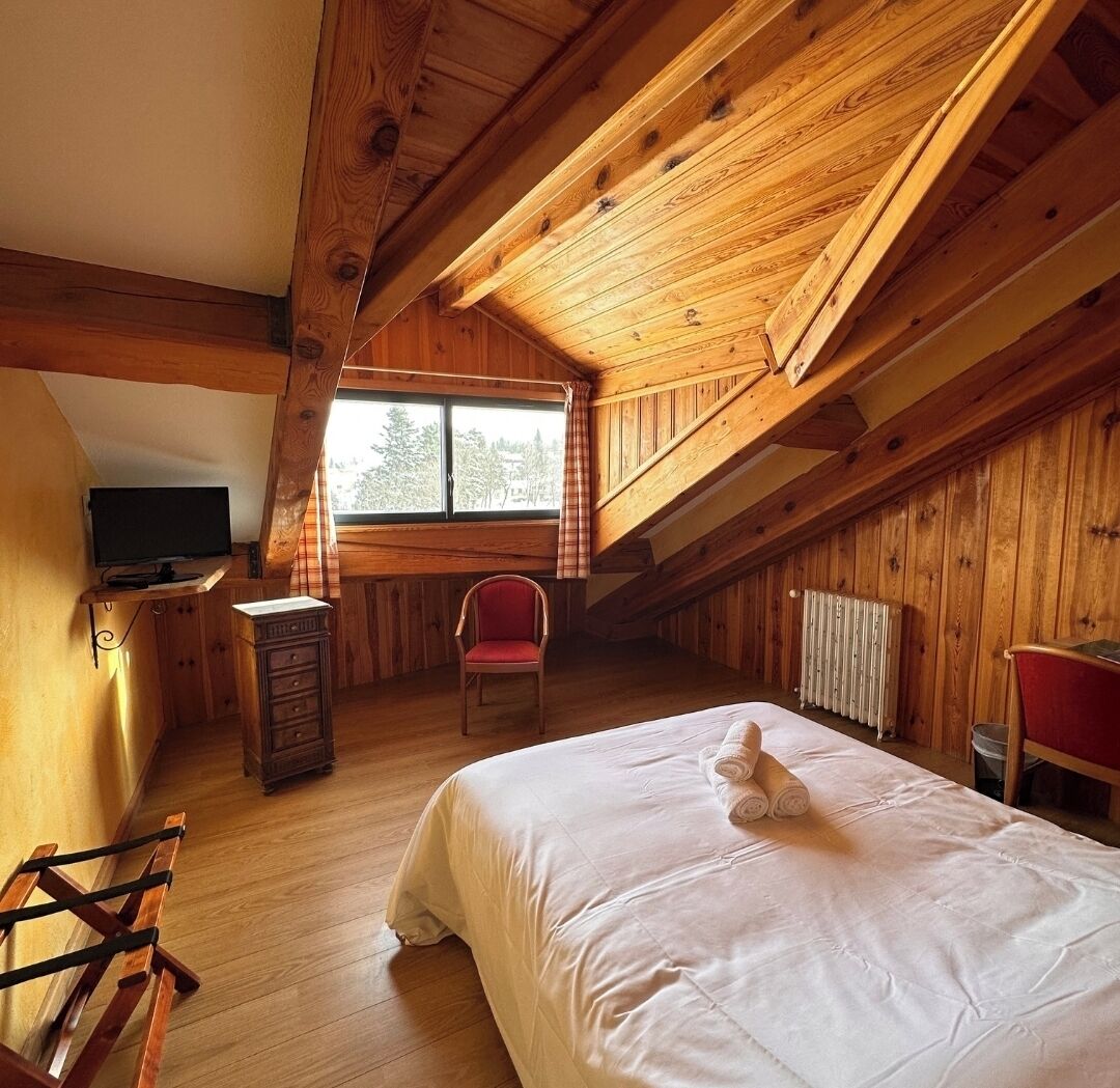 A wooden room with a bed, a chair, a desk with a TV, and a window.