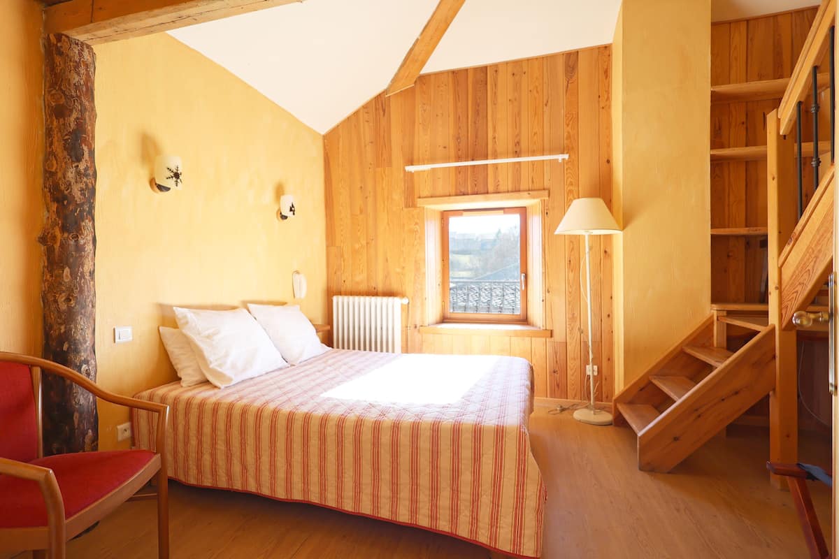 A wooden room with a bed, a chair, a window, and a staircase.