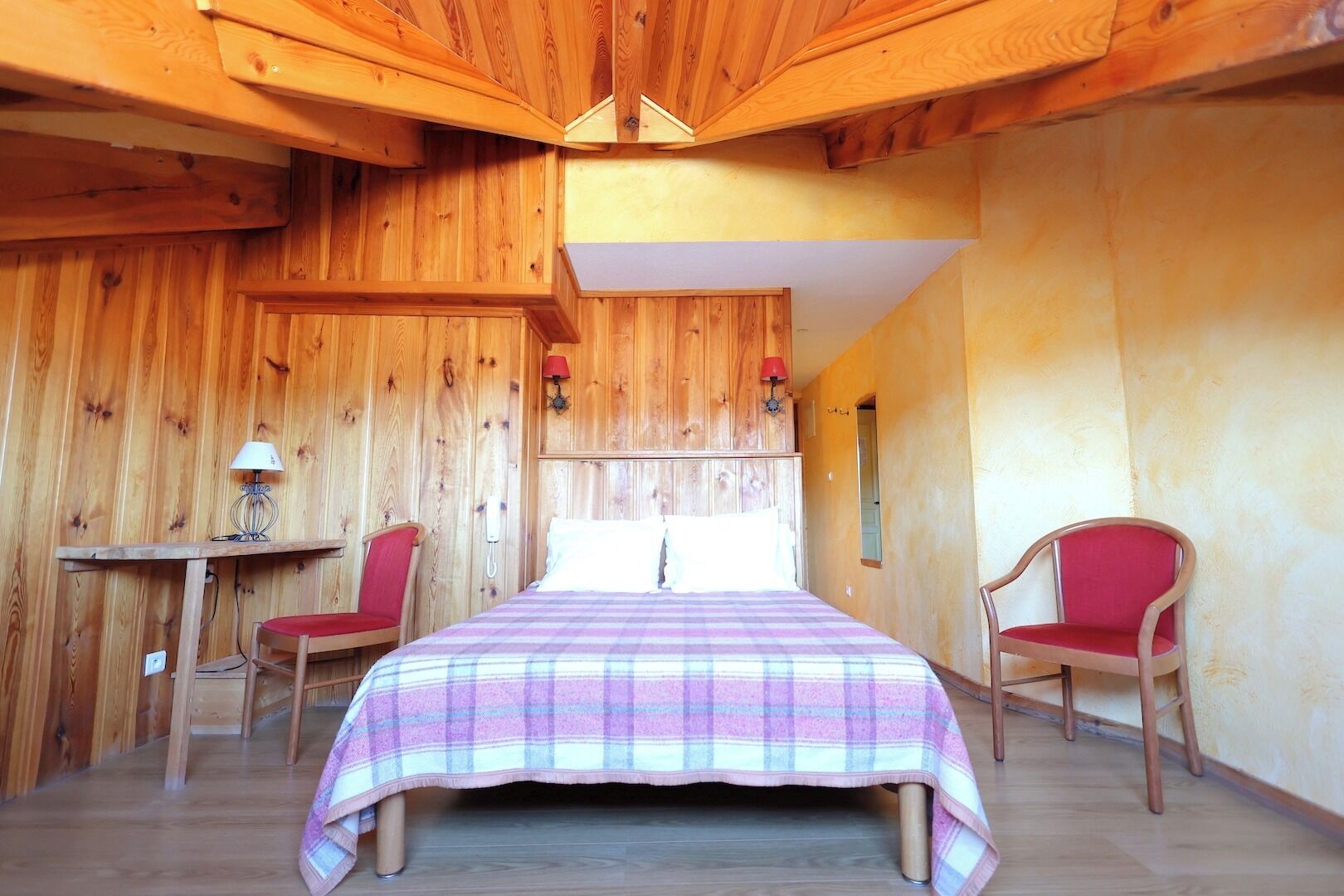 A wooden room with a bed, two chairs, a table, and a lamp.