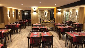 Un restaurant aux murs en bois, agrémenté de pierres apparentes et dont les tables sont recouvertes de nappes rouges. On y trouve des verres à vin et des couverts sur chaque table.