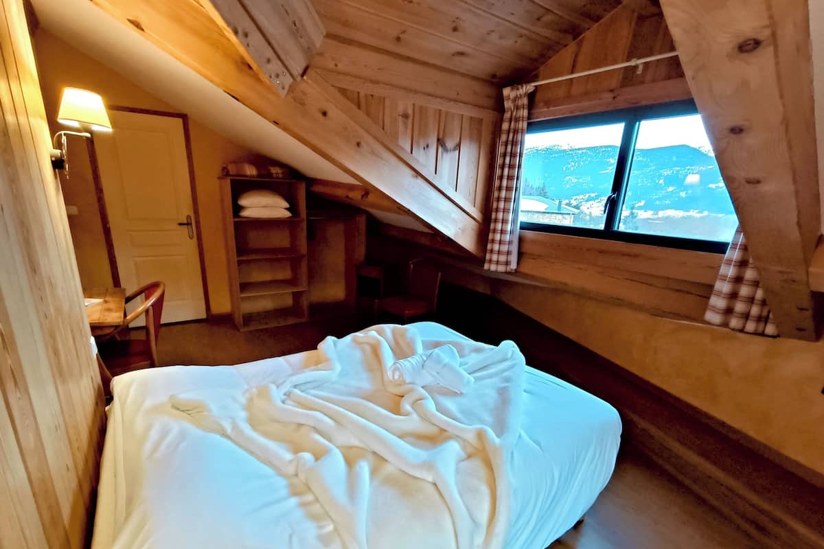 A wooden cabin room with a bed, a chair, a shelf with towels, and a window with a view.
