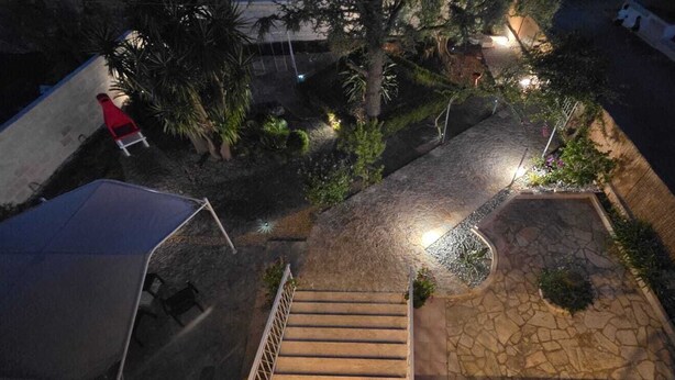 Une vue nocturne d’une cour avec un escalier en pierre menant à une zone abritée, entourée de végétation et agrémentée d’une chaise rouge.